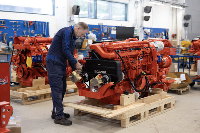 En tekniker jobber med en stor, rød Scania-motor på en pall i et verksted, omgitt av verktøy, maskindeler og flere motorer i bakgrunnen.