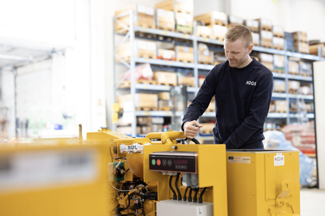 Montør i blå NOGVA-genser jobber med gul strømgenerator i verkstedmiljø, med hyller og reservedeler i bakgrunnen.