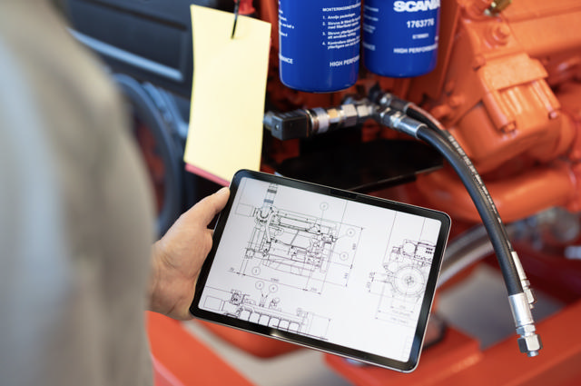 Person holder nettbrett med teknisk tegning av motor foran fysisk motor med hydraulikkslanger og oljefiltre i et verkstedmiljø.