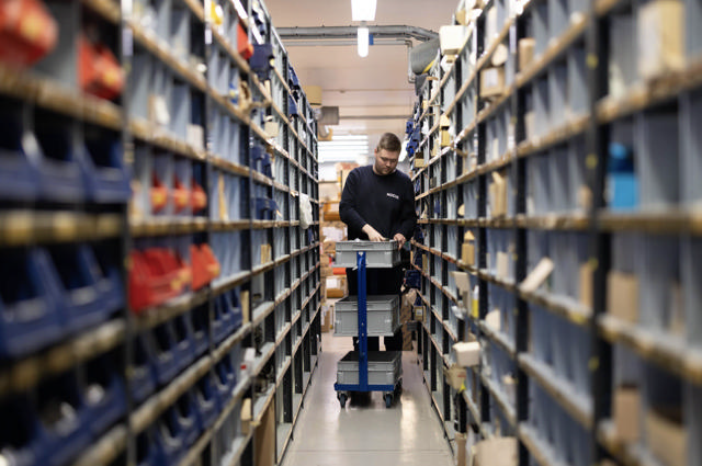 Ansatt triller vogn med plastkasser mellom høye reoler fylt med smådeler og komponenter i et lagerrom hos Nogva.